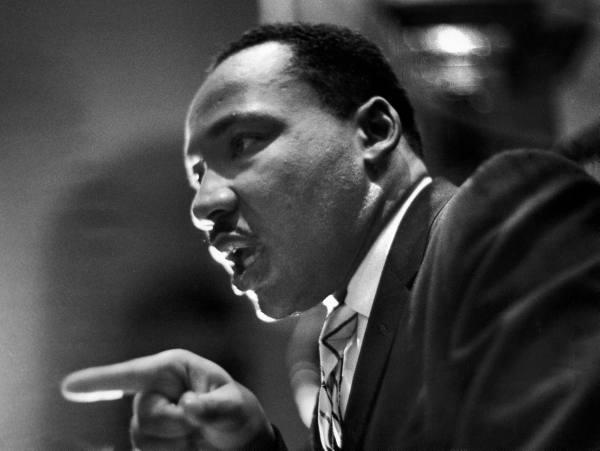Martin Luther King, Jr. speaks in a church. He leans forward, eyes gleaming with intensity, a finger pointed with conviction out at his audience.