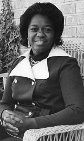 Juanita W. Goggins, an African-American woman, sits in a wicker chair that is seemingly positioned on a porch. The photo is in black and white. Goggins wears a dark jacket with a tie around the waist, two sets of white buttons down the front, and large white lapels. Her hands are folded neatly in her lap. She looks directly at the camera and smiles.
