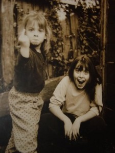 Two fair-skinned little girls.  One is flipping off the camera with a huge frown, and the other is laughing at the other's gumption.  Both look fabulously naughty.