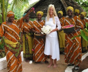 White woman barefoot in a white Western-style wedding dress surrounded by African women in traditional dresses. 