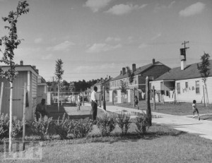 black and white image of sorjourner truth homes in Detroit Michigan. THere are two rows of homes with a long sidewalk down the middle. the homes are small but neat single family homes. there is a black man outside--it's hard to tell what he is doing.