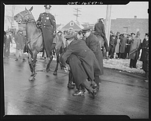 a protestor struggles with two white police officers. One police officer has his arm around the protestor as if to throw the person down. A white cop on a horse watches.