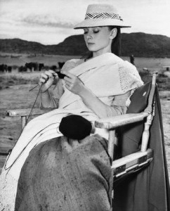 Audrey Hepburn sits in a folding chair in the middle of an empty plain, wearing period costume and knitting