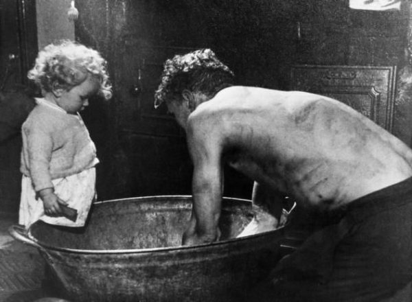 Grainy, black-and-white historical photo of a man leaning over a galvanized bathtub as a child looks on