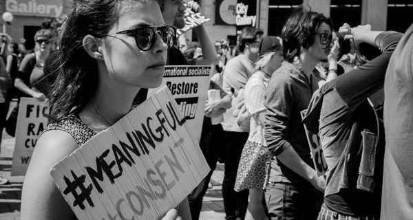 A college student protests with a cardboard sign reading "#MeaningfulConsent"