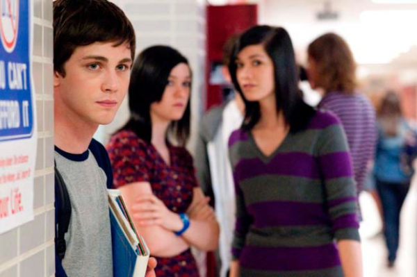 Movie still from "The Perks of Being a Wallflower," set in a high school, with a young man in the foreground looking off into the distance as two young women stand in the near background, talking about him