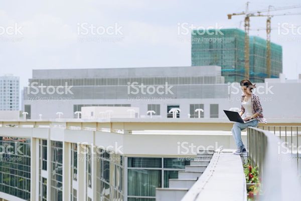 Young woman in jeans and plaid shirt uses laptop while sitting on the edge of a rooftop